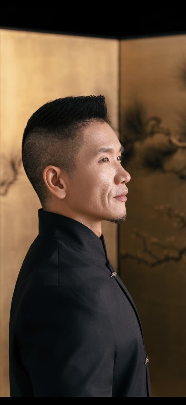 Hao Pan in profile wearing a dark mandarin-collar jacket against a gold folding screen